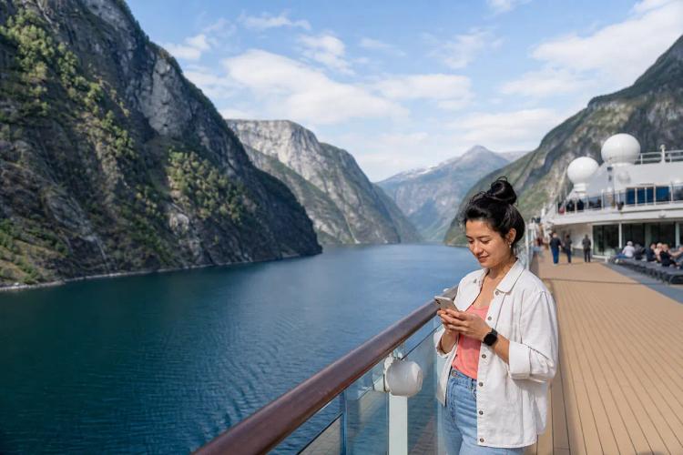 Female passenger checking her phone at the railing of a ship sailing through a Norwegian fjord