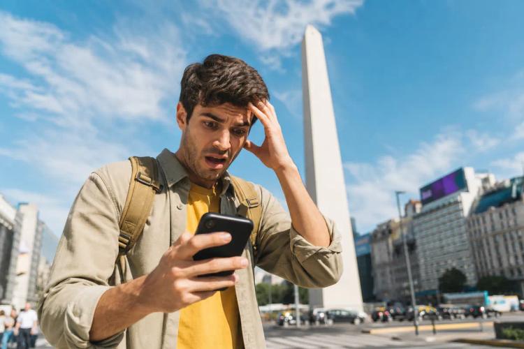 Voyageur atterré regardant son téléphone, main dans les cheveux, devant un obélisque en plein soleil