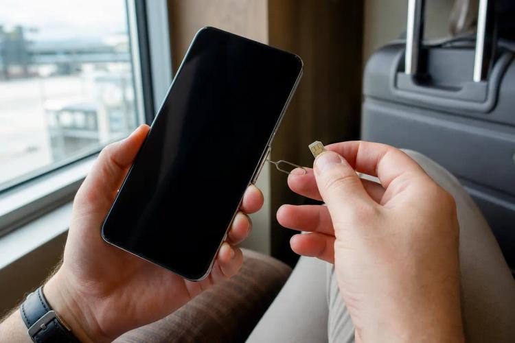 Close-up of hands holding a SIM card and ejector pin next to a switched-off smartphone