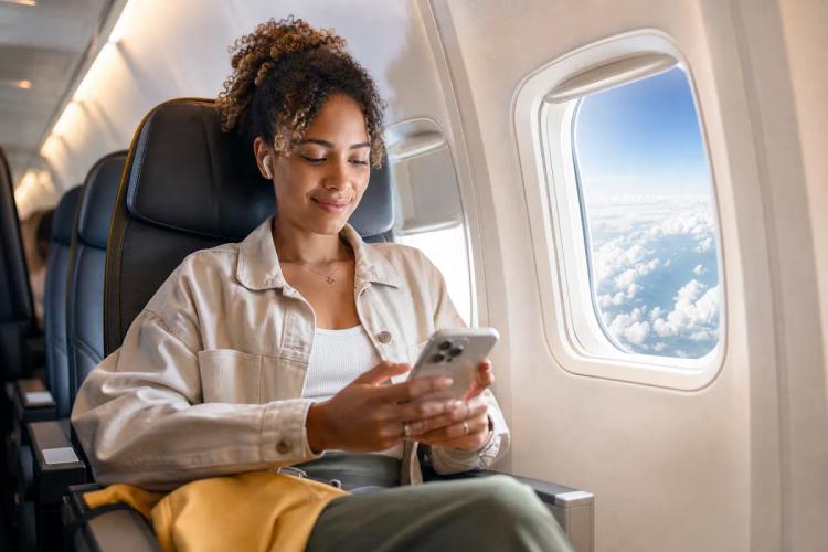 Relaxed passenger using her smartphone beside an aircraft window with clouds below