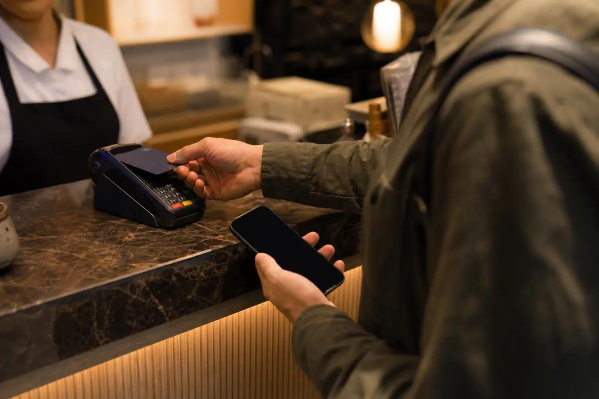 Customer tapping a bank card on a payment terminal at a cafe counter, phone in the other hand
