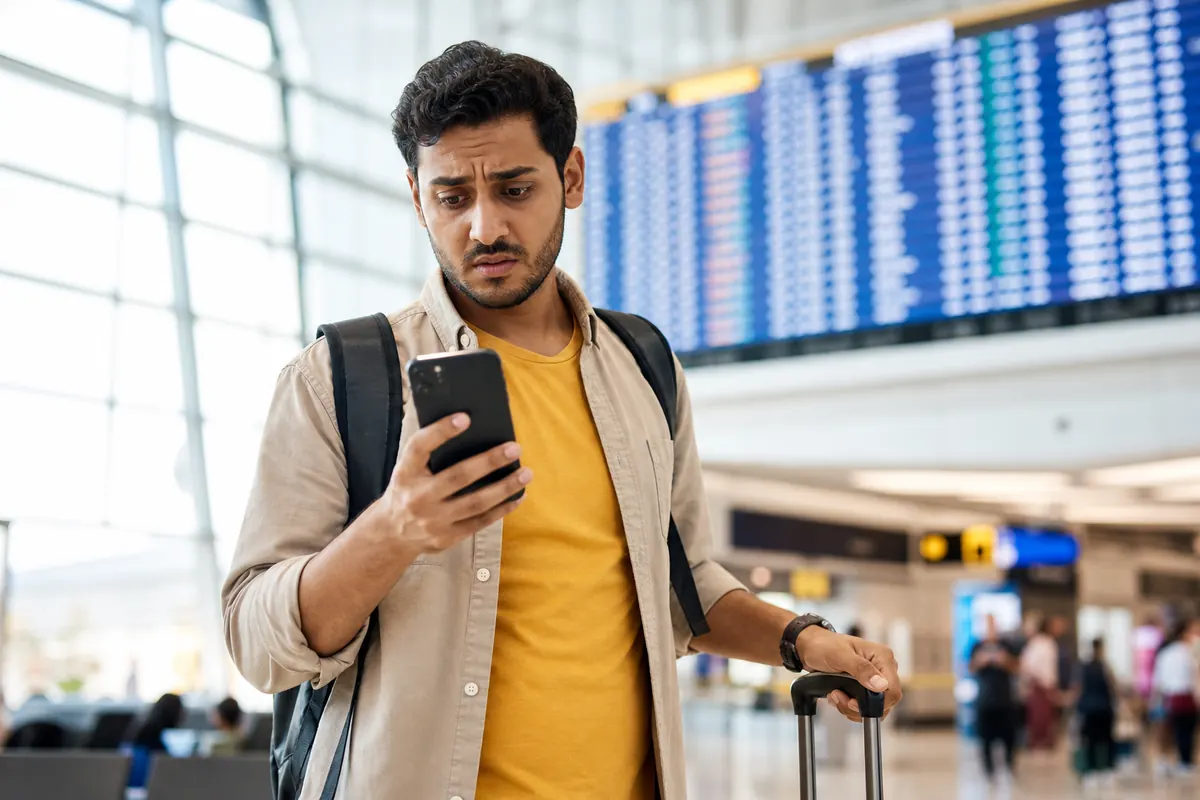 Worried traveler checking their smartphone in front of an airport flight departure board.
